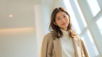 Young Asian businesswoman in beige coat standing confidently in modern office building with natural lighting for corporate and professional concepts.