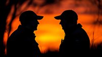 Two men in baseball caps silhouetted against dramatic orange sunset sky, sharing conversation outdoors in nature, dark profile shapes create striking contrast.