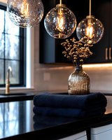 Modern kitchen interior with glass pendant lights illuminating dark countertop, featuring folded navy towels and decorative vase with dried flowers. Elegant home decor.