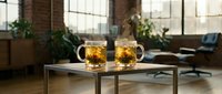 Two glass mugs filled with golden tea on modern coffee table in bright loft office space with brick walls and large windows for cozy workplace atmosphere.