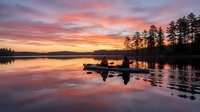 Serene lake at sunrise with golden and pink sky reflecting on calm water, silhouetted trees and rocks creating tranquil nature landscape for backgrounds and meditation content.