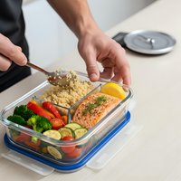 Hands preparing healthy meal prep lunch box with salmon, quinoa and fresh vegetables in glass container for balanced nutrition and diet planning.