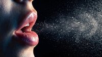 Close-up of woman's lips exhaling fine mist or spray particles against dark background, creating dramatic lighting effect for health and beauty concepts.