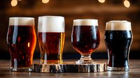 Four different craft beers in distinctive glasses on wooden coaster, showing variety from light lager to dark stout with rich foam heads against warm bokeh background.