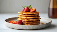 Stack of golden pancakes topped with fresh strawberries and maple syrup on gray ceramic plate, morning breakfast setting with natural light.