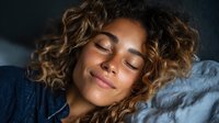 Young woman with curly hair and freckles sleeping peacefully on pillow, illuminated by soft morning light against dark background.