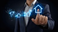 Professional in dark suit touching glowing blue house icon with rising graph trend, representing digital real estate market growth and property investment technology.