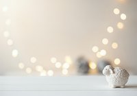 Cute white ceramic sheep figurine on wooden surface with warm golden bokeh lights and blurred ornaments in background, perfect for holiday greeting cards, seasonal marketing, and festive home decor projects.