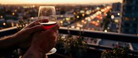 Hand holding wine glass with red wine against blurred city lights at sunset, rooftop celebration and evening relaxation concept for hospitality and lifestyle.