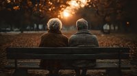 Elderly couple sitting together on park bench during golden hour sunset with autumn leaves scattered around creating romantic peaceful moment.
