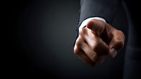 Close-up of businessman's hand pointing directly at viewer against dark background, conveying authority and accountability in professional setting.