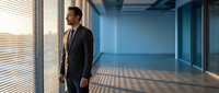 Professional businessman in dark suit standing by office windows with sunlight streaming through blinds creating dramatic shadows on modern corporate floor.