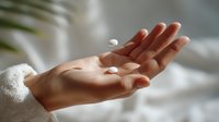Open palm of a woman in white sweater holding two white oval pills against soft blurred background, representing medication or supplement use.