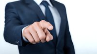 Businessman in navy suit pointing finger forward against white background, conveying leadership and decision-making in corporate environment.