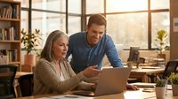 Mature woman and middle-aged man collaborating at laptop in bright office with large windows, wooden shelves, and plants. Suitable for business, teamwork, and professional training content.