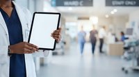 Doctor in white coat and stethoscope holding blank tablet in modern hospital corridor with blurred medical staff and patients in background, suitable for healthcare technology and medical services.