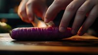 Purple carrot being sliced on wooden cutting board in dramatic lighting, close-up view showing knife skills and food preparation in kitchen setting.