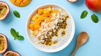Overhead view of a white bowl filled with yogurt, granola, mango chunks, and passion fruit seeds on a bright blue surface with fresh fruit and mint leaves.