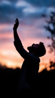 Silhouette of young man with raised arms against dramatic sunset sky, expressing freedom, spirituality and worship. Moody dusk atmosphere with pink and blue clouds.