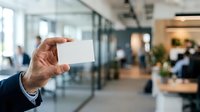 Businessman in dark suit holding blank white business card in modern office environment with glass partitions and colleagues in background.