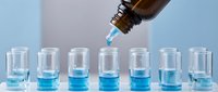 Laboratory technician dispensing blue chemical solution from amber bottle into test tubes for scientific research and pharmaceutical analysis.