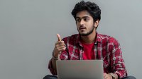 Young South Asian man in red plaid shirt and t-shirt gesturing while working on laptop against gray background, conveying positive communication during online meeting.
