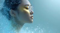 Young Asian woman with glowing golden makeup and glasses in profile view against turquoise background with magical sparkling particle effect dissolving into space.