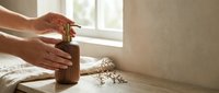 Woman hands using wooden pepper mill grinder on kitchen counter with scattered peppercorns near window creating warm natural lighting atmosphere.