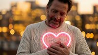 Middle-aged Caucasian man in cream sweater holding glowing pink neon heart at sunset with warm city bokeh lights creating romantic atmosphere for love concepts.
