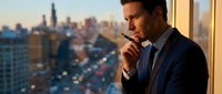 Professional businessman in suit smoking cigarette by office window overlooking city skyline at sunset. Corporate executive contemplating business decisions.