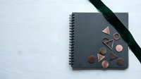 Gray spiral notebook with geometric copper shapes and green ribbon on white marble surface for creative design projects and artistic planning.