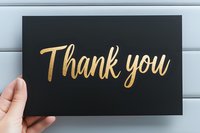 Hand holding black card with elegant gold metallic thank you lettering against light wooden background. Minimalist gratitude message for special occasions.