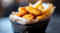 Crispy golden chicken nuggets served in wire basket with french fries on white paper liner, close-up view showing texture and warm tones of fried food.