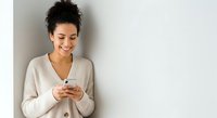 Young woman with dark curly hair and medium skin tone wearing beige cardigan, smiling while holding smartphone against white wall background for business and lifestyle content.