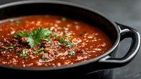 Homemade tomato soup garnished with fresh herbs and ground pepper in black cast iron pot, close up view showing rich texture and vibrant color.