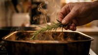 Hand holding fresh rosemary sprig over steaming pot with roasted meat, warm kitchen lighting, cooking preparation and culinary ingredient concept.