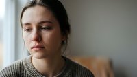 Young Caucasian woman with brown hair looking sad and contemplative by window, expressing depression, loneliness, and mental health struggles indoors.