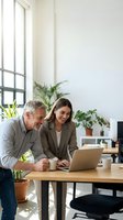 Mature man and young woman working together at wooden desk with laptop in bright office space with plants and natural light from window.