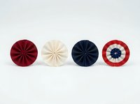 Colorful pleated fabric rosettes in red, cream, navy and red-white stripes arranged in a row on white background, isolated design elements for crafts and decorative projects.