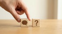 Hand placing wooden cube with question mark next to block with speech bubble symbol on desk representing communication and inquiry concepts.