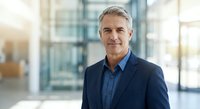 Confident mature businessman with gray hair in navy suit standing in modern office building lobby with natural lighting for corporate presentations.