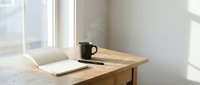 Wooden desk with open notebook and black coffee mug by bright window, minimalist workspace setup for creative work, writing, or remote office environment.