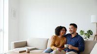 Young couple, mixed ethnicity, sitting together on beige sofa in bright modern living room, holding tablet device, smiling and engaged in conversation about digital content or online shopping.