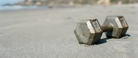 Weathered metal dumbbells resting on sandy beach with blurred coastal background, representing fitness, strength training, and outdoor workout equipment for health and wellness concepts.