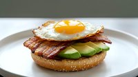 Gourmet breakfast sandwich with fried egg, crispy bacon and fresh avocado slices on sesame seed bun served on white plate for healthy morning meal concepts.