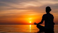 Woman meditating in lotus pose silhouetted against golden sunset over calm ocean water creating peaceful zen atmosphere for wellness and mindfulness practice.