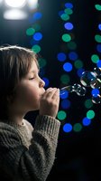 Young child blowing soap bubbles against colorful bokeh lights background. Caucasian kid in knit sweater creating magical moment with bubble wand for childhood memories.