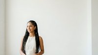Young woman with long braided hair and dark skin tone wearing a white sleeveless top, smiling while looking to the side against a neutral white background for portrait and lifestyle use.