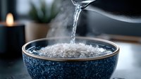 Pouring hot water into blue ceramic bowl with rice grains creates rising steam in moody lighting, candle and plant in background for food photography.