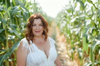 Curvy Caucasian bride with auburn wavy hair and natural makeup poses confidently in white lace wedding dress among tall green corn stalks on sunny summer day.